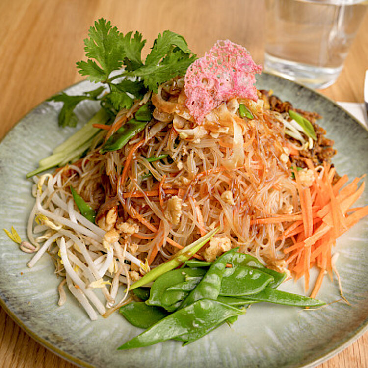 Ein Teller mit einem bunten asiatischen Nudelgericht, garniert mit frischen Gemüsestreifen, Sprossen, Erdnüssen, Koriander und einem knusprigen roten Chip. Im Hintergrund steht ein Glas Wasser, daneben liegen Besteck und eine Serviette.