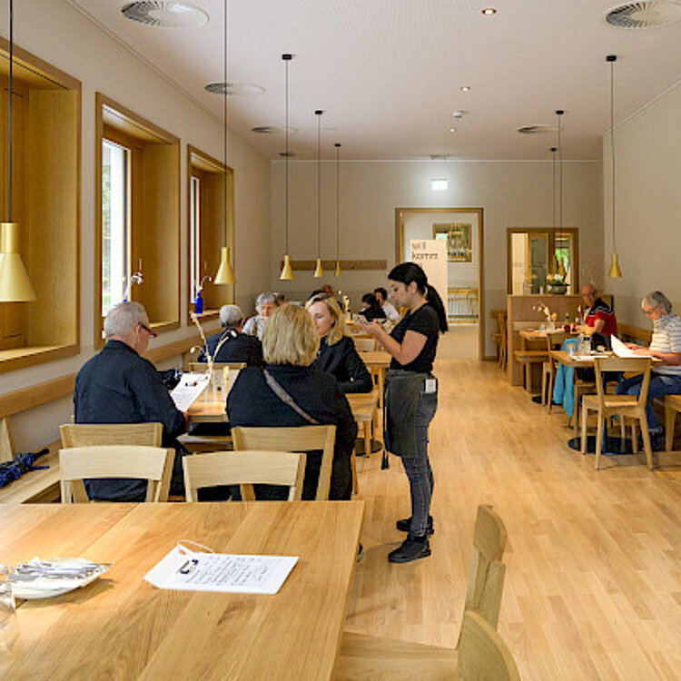 Eine moderne Cafeteria mit mehreren Gästen an Holztischen, eine Kellnerin bedient die Gäste, helle und freundliche Atmosphäre mit großen Fenstern und Holzfußboden.