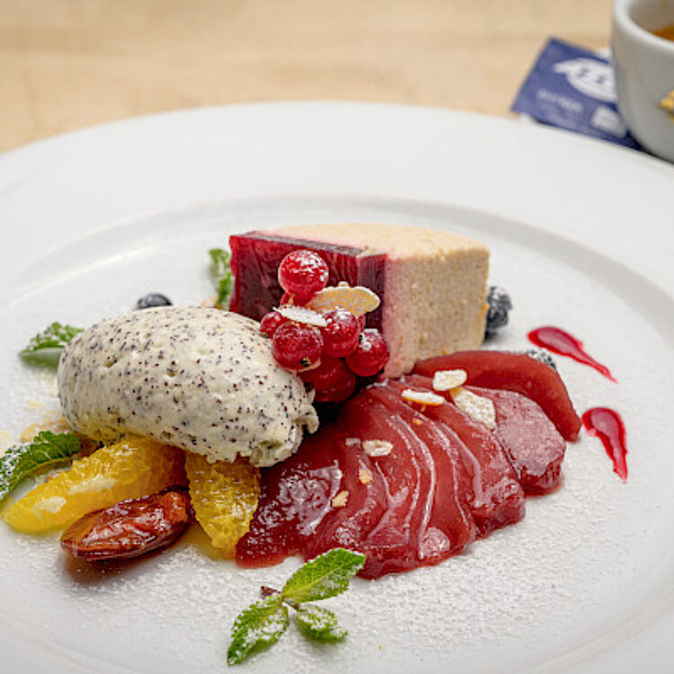 Ein kunstvoll angerichtetes Dessert mit einer Schicht aus roten Beeren, einer Kugel Mohn-Eis, Orangenscheiben und frischen Minzblättern auf einem weißen Teller, garniert mit Puderzucker. Im Hintergrund steht eine Tasse Espresso.