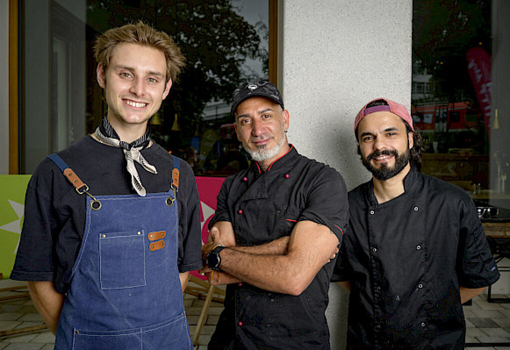 Drei männliche Köche in Arbeitskleidung, lächelnd, stehen zusammen vor einem Fenster.