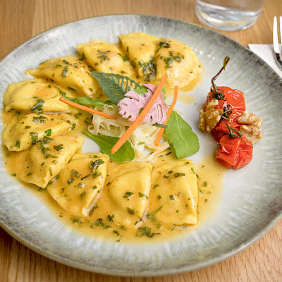 Ein Teller mit Ravioli in hellgelber Sauce, garniert mit Kräutern, Salatblättern und gerösteten Kirschtomaten, daneben Besteck auf einer Serviette.