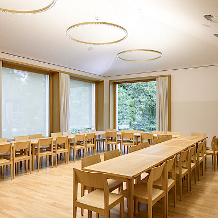 Ein heller, moderner Konferenzraum mit großen Fenstern, zwei langen Holztischen und mehreren Holzstühlen, die in Reihen nebeneinanderstehen. Die Decke ist weiß mit runden Lichtquellen ausgestattet.