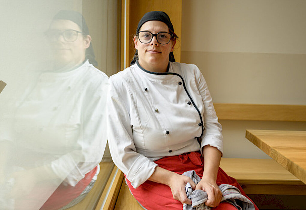Porträt einer Köchin in weißer Kochjacke, roter Schürze und schwarzer Mütze, die entspannt auf einer Holzbank neben einem Fenster sitzt und in die Kamera blickt.