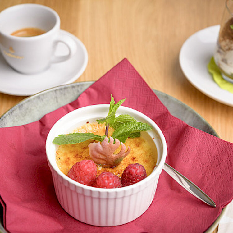 Eine Creme Brûlée in einer weißen Schale, garniert mit Himbeeren, Minzblatt und einer Schokoladendekoration, auf einem roten Servietten-Teller. Im Hintergrund eine Tasse Kaffee und ein Dessertgläschen mit Schichtdessert.
