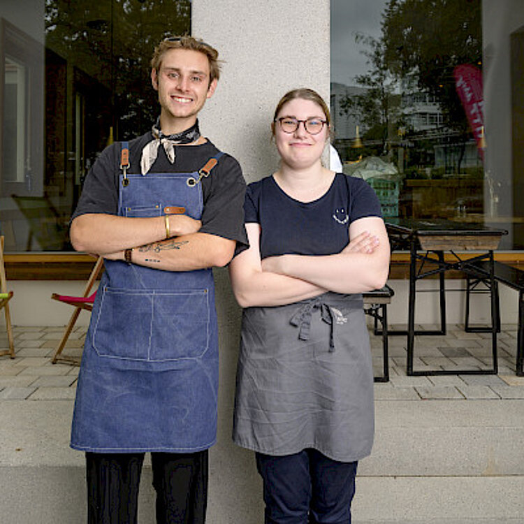 Zwei junge Gastronomiemitarbeitende stehen lächelnd mit verschränkten Armen vor einem Café-Eingang, beide tragen Schürzen und wirken freundlich.