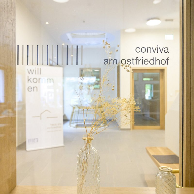 Blick durch eine Glastür in einen hellen, modernen Eingangsbereich mit einem Schild „conviva am ostfriedhof“ und einem Willkommensplakat im Hintergrund. Im Vordergrund steht eine Vase mit getrockneten Blumen auf einer Holzoberfläche.
