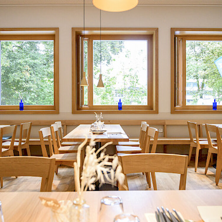 Helles, modernes Restaurant mit Holzmöbeln, großen Fenstern und natürlichem Tageslicht, das eine ruhige und einladende Atmosphäre schafft.