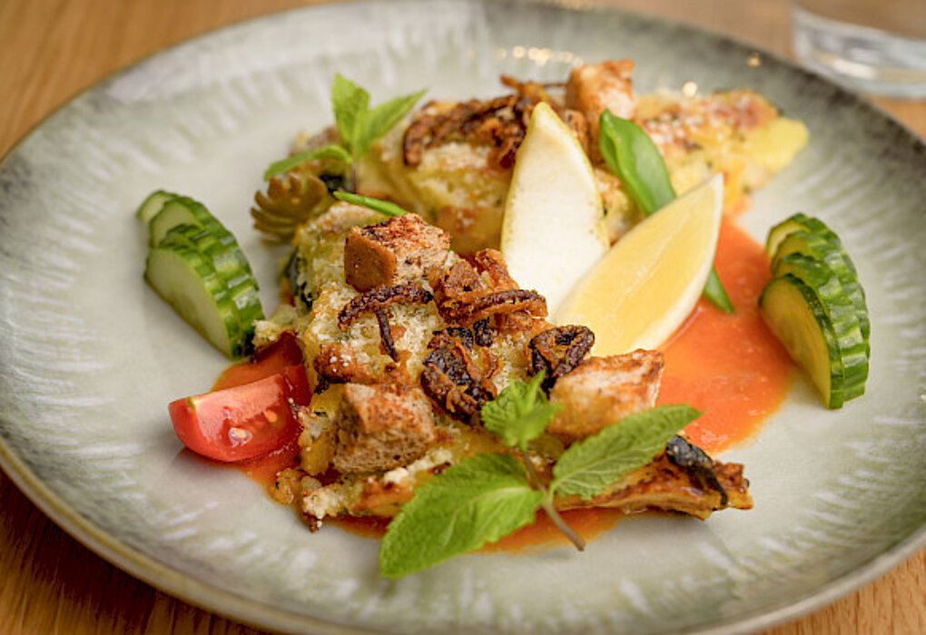 Ein Teller mit einem Gericht aus gebackenem Fisch, garniert mit Zitronenspalten, Tomatenstücken, Gurkenscheiben, frischen Kräutern und einer roten Sauce.